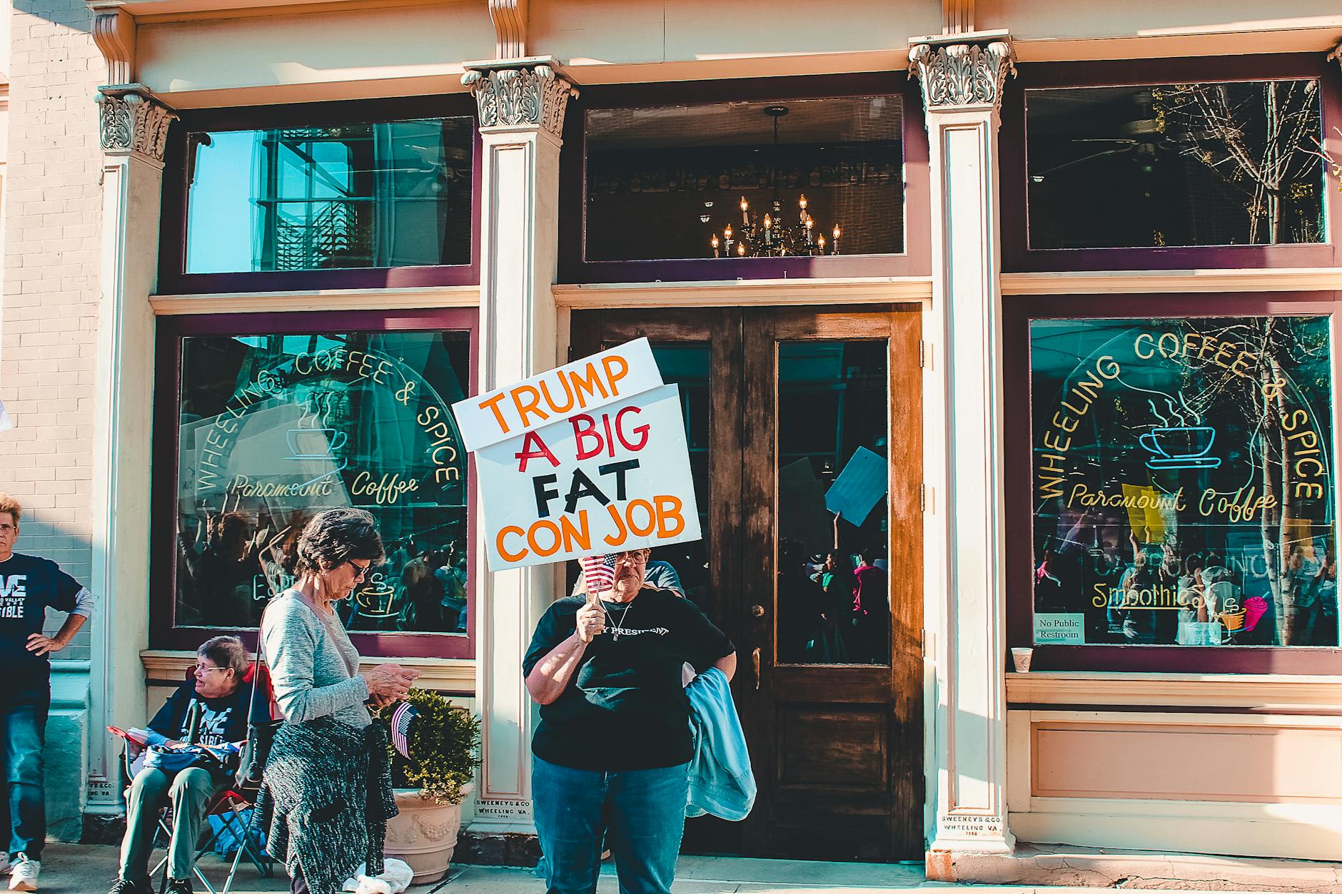 Protesters express political opinions in front of coffee shop with vibrant signs against Trump.