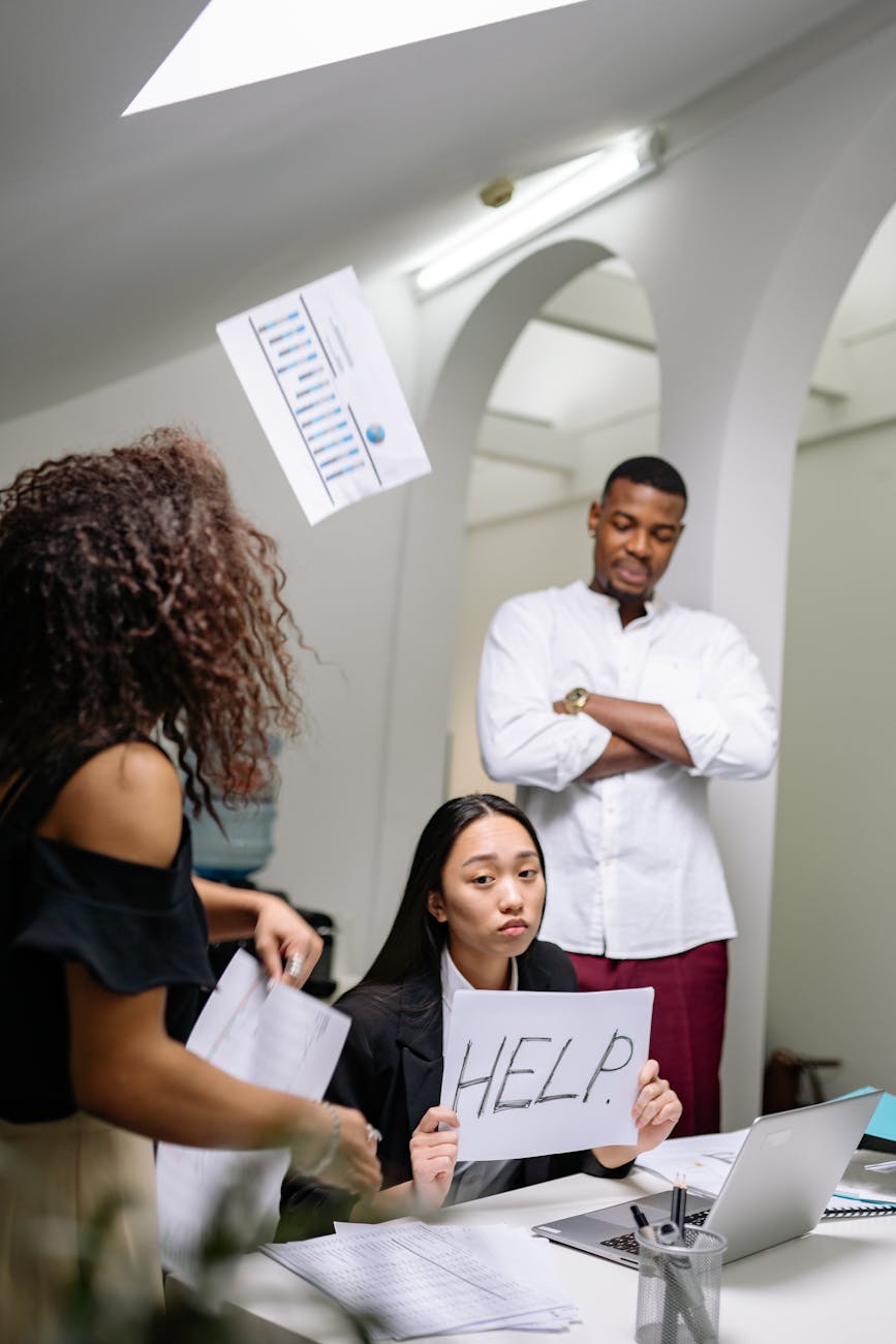 Woman holding 'HELP' sign with coworkers around, depicting workplace stress.