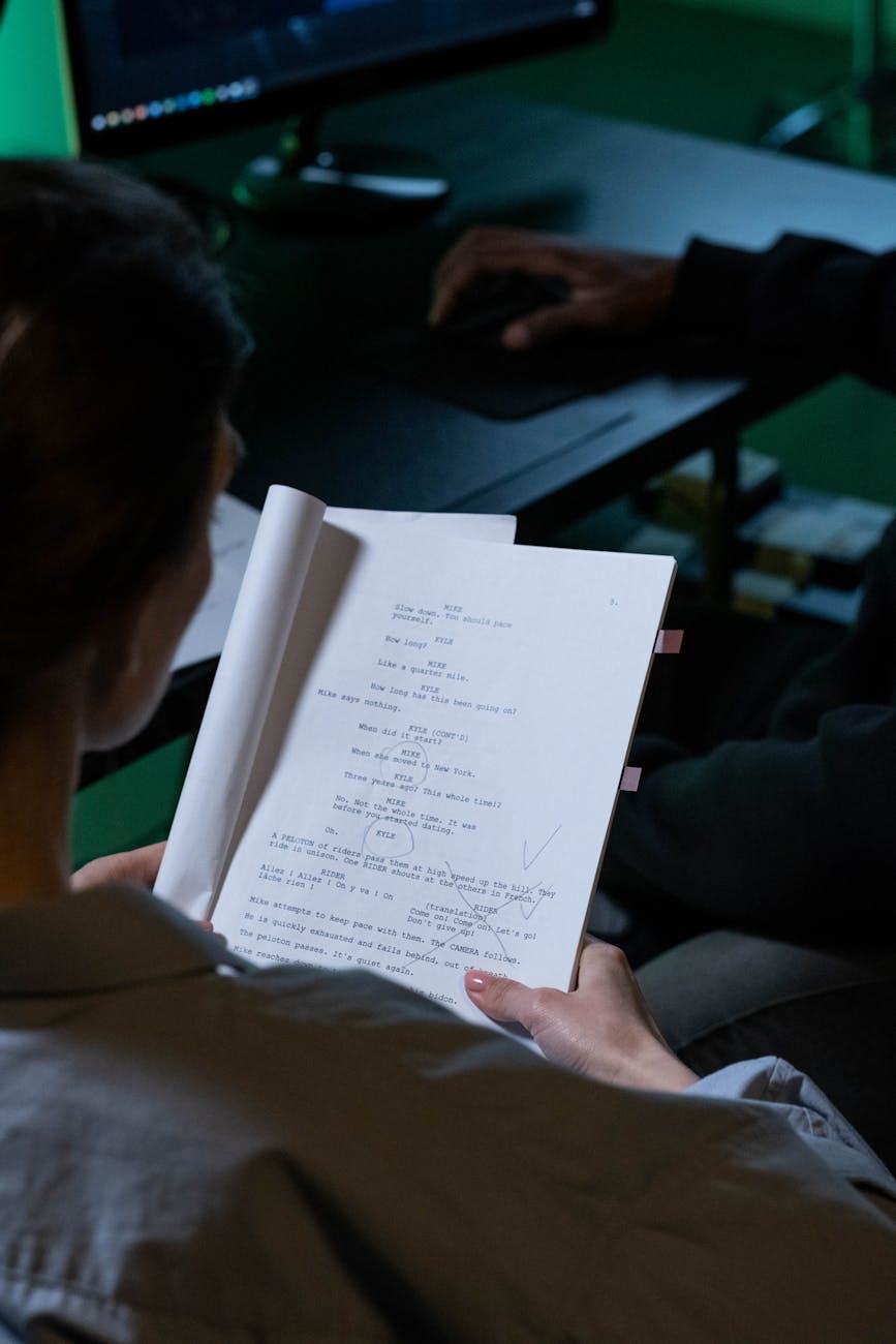 Person reading a script with notes in a dimly lit workspace, viewed over the shoulder.