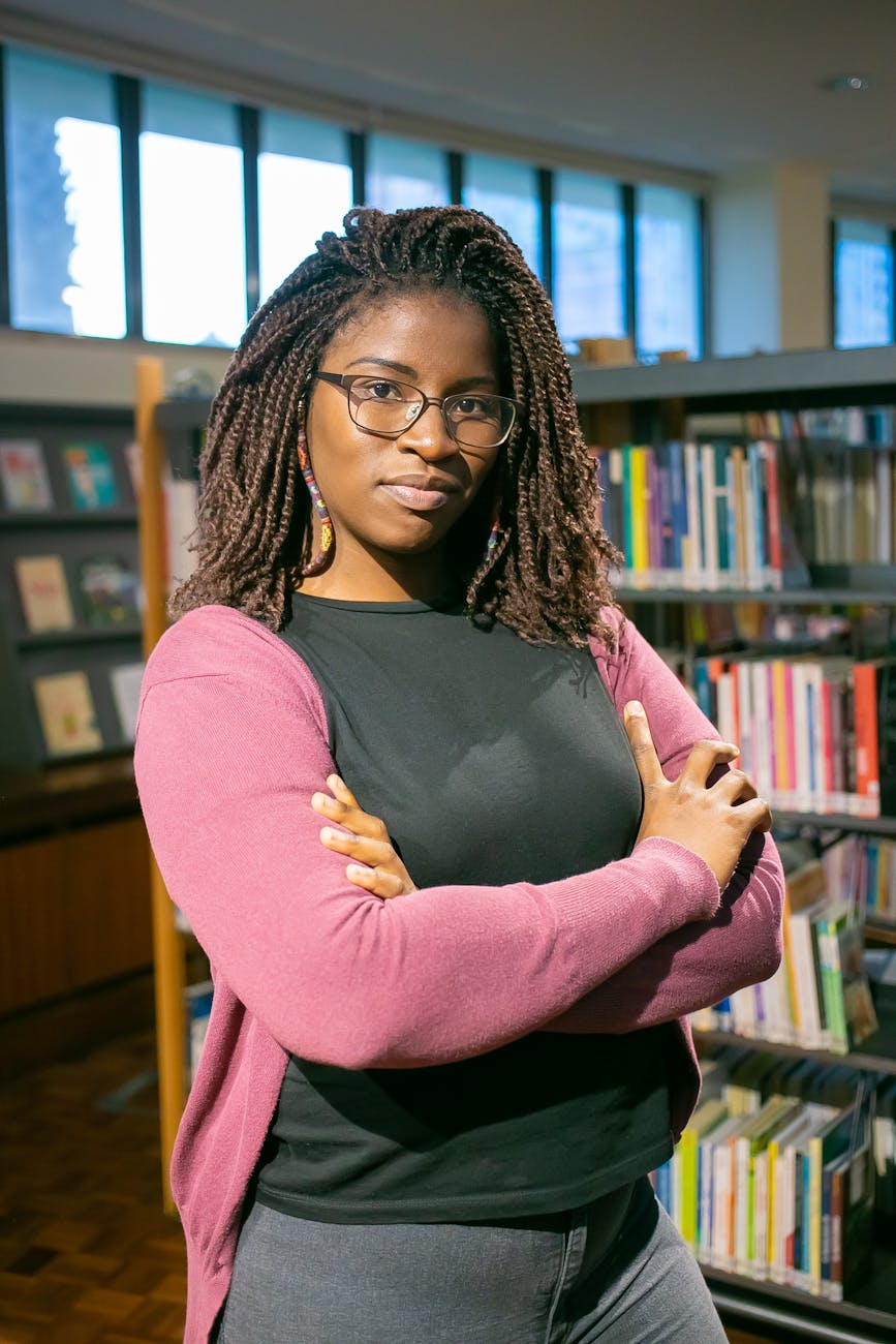 Young woman with glasses stands confidently with arms crossed in a library.